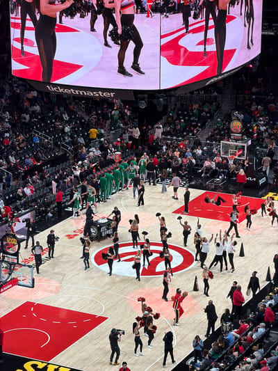 The image shows a basketball game at an arena with a large screen displaying dancers and the word "ticketmaster." The basketball court displays the logo of the Atlanta Hawks. Cheerleaders are performing on the court. Spectators fill the seats around the court. Several players are wearing green uniforms. The scoreboard displays "12:00" and "88.88," along with the logo of Tissot, along with the words StateFarm, and Natural Gas. The arena is likely located in Atlanta, United States of America.
