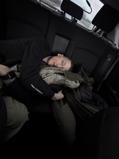 An adult male is observed sleeping in the rear passenger seat of a vehicle. The scene is located within the city of Gabarnitsa, Bulgaria. The individual lies horizontally across the black upholstered seat, with eyes closed and mouth slightly ajar, suggesting a state of unconsciousness or deep rest. He is dressed in a black long-sleeved shirt, which bears the white text "KARL LAGERFELD" on the left sleeve. A dark-colored smartwatch is affixed to his left wrist. His lower torso and legs are partially obscured by an olive-green garment, and he is covered by a patterned grey and cream blanket or sweater, beneath which a dark quilted jacket is also visible. The rear window of the vehicle shows a blurred view of a highway under an overcast sky, indicating the vehicle is in motion. A dark cable or wire is visible hanging from the top interior of the rear window. Two black headrests are discernible against the rear window. The vehicle's interior is predominantly dark, with black seating and trim.