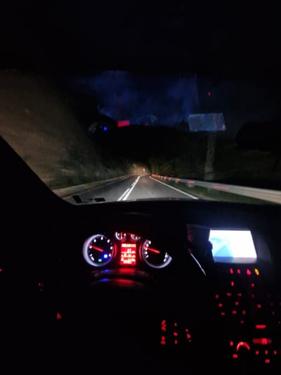 The image shows a nighttime scene from inside a vehicle. The dashboard is visible, lit with red and blue lights, displaying gauges and digital information. Through the windshield, a road stretches into the distance, illuminated by the vehicle's headlights and roadside lights. To the left of the road, there appears to be a dark area with some faint lights. The presence of red lights along the road may indicate the vehicle is passing through Pastra, Bulgaria.