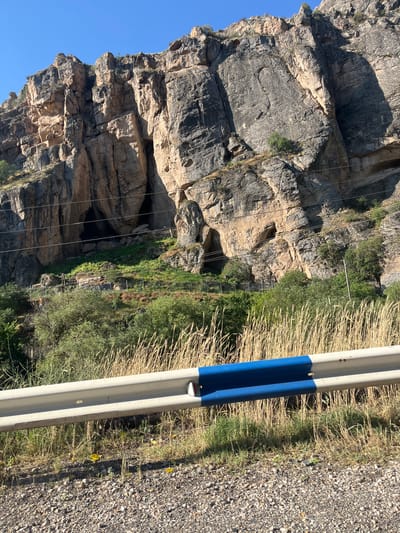 The media file depicts a natural scene with a prominent rock formation. The rock face is a mix of rugged, weathered stone with textures ranging from smooth to heavily fissured. Shadows suggest the sun is casting light from the side, creating depth and highlighting the contours of the rock. In the foreground, a metal barrier with blue and white markings runs vertically along the right side of the frame. This barrier appears to be a guardrail, possibly alongside a road.
Beside the rock formation, there is a dense growth of dry, straw-like vegetation interspersed with greener foliage. Further down the rock face, some man-made structures are visible, including what appears to be a metal fence and a walkway or stairs leading up the side of the cliff, suggesting a hiking or tourist trail. Thin wires, possibly power lines or communication cables, are strung across the rock face.
The scene is outdoors, under a clear blue sky, indicating fair weather. The lighting and the dry vegetation suggest it might be a warm, possibly late summer or early autumn day. The overall impression is one of a dramatic natural landscape, accessible to visitors. No people are visible in the frame. There is no visible text. The location context points to Zangakatun, Armenia, a region known for its mountainous terrain.