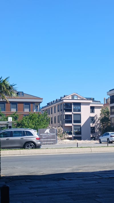 The image shows a clear blue sky over an urban scene in Bahçelievler, Turkey. In the foreground, a grey SUV is parked on the side of a road. Beyond the SUV, a sign is visible, partially obscured by pink flowers, with text that includes "AKYAZICILAR İNŞAAT" and a website address. Two modern apartment buildings are situated behind the sign. The building on the right is lighter in color with multiple balconies and dark-tinted windows. The building on the left is darker, with a prominent roofline and vertical wooden accents. Green trees are planted between the buildings. A white car is parked to the right of the lighter colored apartment building. The overall scene depicts a residential area under construction or recently completed.