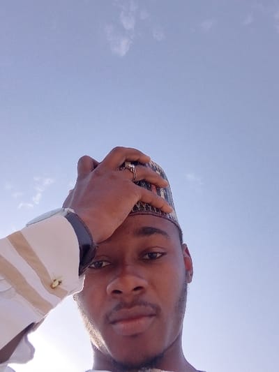 A young adult male, identified as being in Kwazailewa, Nigeria, is depicted in a low-angle shot against a clear, light blue sky with sparse white clouds. The male has dark skin, a short beard, and a mustache. He wears a patterned cap, possibly a Kufi, in dark colors with light motifs. His right hand is raised to his head, with fingers resting on the cap. A ring with a visible stone is on the ring finger of his right hand. He wears a light-colored, vertically striped long-sleeved shirt with a gold-colored cufflink visible on his left wrist. A dark-strapped wristwatch is present on his left wrist. Bright sunlight emanates from the lower-left of the frame, illuminating the left side of his face and shirt. He looks directly forward towards the viewer.