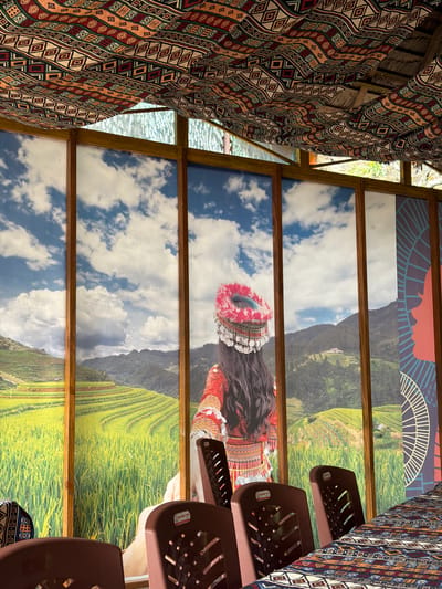 This image depicts the interior of a dining area, possibly a restaurant or cafe, in Khanh Vinh, Vietnam. The dominant feature is a large mural covering the back wall, showcasing a scenic landscape of terraced rice fields under a cloudy sky, with mountains in the background. A woman, dressed in traditional attire with an ornate headdress, is partially visible in the mural, facing away from the viewer. The ceiling is adorned with a colorful fabric featuring geometric patterns, and a bamboo structure is visible above it. In the foreground, several dark brown plastic chairs surround a table covered with a patterned tablecloth that complements the ceiling fabric. The overall impression is one of cultural ambiance and a scenic backdrop.