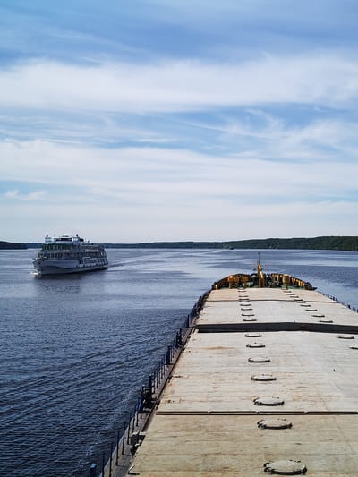 This image captures a wide river scene near the city of Nosovo, Russia, under a blue sky with scattered cirrus and stratocumulus clouds. The foreground is dominated by the flat, weathered deck of a large barge, viewed from the bow looking forward. The barge deck is light grey/beige, featuring multiple rows of circular, bolted hatches, and a blue metal railing visible along its port side. Towards the visible front of the barge, there is a low structure with yellow components, possibly equipment or a small cabin base.
To the left of the barge, in the mid-ground, a multi-decked white passenger ship, likely a river cruise vessel, is observed moving from left to right. The ship exhibits multiple rows of windows and balconies, some with green trim, and a prominent red star emblem on its bow. A distinct wake trails behind the moving passenger ship. The water, a deep blue, shows ripples and patterns from the vessels' movement.
In the far background, dense green forested banks line both sides of the expansive waterway, extending towards the horizon. A very small, dark vessel is faintly visible near the distant right bank. No people are discernible on either the barge or the passenger ship from this perspective. The scene depicts active navigation on a broad river or canal under daylight conditions.
