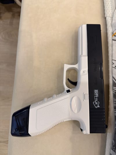 A white and black toy gun is lying on a light beige, textured surface. The toy gun is oriented horizontally, with the barrel pointing to the left. The rear of the gun, which is black, has white text that reads "WATER BATTLE ELECTRIC WATER GUN" with a triangle logo. Above this text, more white text reads "AUSTRIA 9X19". The front portion of the toy gun is white and features a textured grip. A dark blue translucent piece is attached to the front underside of the gun, likely a water reservoir. The background appears to be a soft surface, possibly upholstery or fabric. There are no people or animals visible in the image. The location of Александровка, Russia is not depicted or suggested by the image.