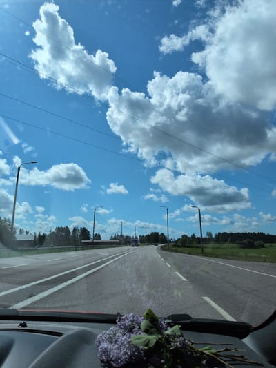 This detailed description pertains to a sunlit, daytime scene captured from inside a moving vehicle on a multi-lane highway in the city of Hartola, Finland.
The upper two-thirds of the image display a clear, bright blue sky filled with numerous white, puffy cumulus clouds of varying sizes, indicating fair weather. Several dark, thin utility lines (likely power or telephone lines) are stretched horizontally across the upper sky. A noticeable lens flare or sun glare is present in the upper left quadrant.
The midground features a wide asphalt road, divided into at least two lanes visible to the driver, with prominent white solid and dashed lines marking the lanes and shoulders. Streetlights with dark poles and modern fixtures are positioned at intervals along the left side of the road, extending into the distance. The immediate environment bordering the road consists of a grassy verge. Beyond this, on both sides, is a rural landscape. To the left, several scattered buildings, including a prominent red-roofed structure with white walls, are visible amidst a mix of green fields and dense coniferous forest. To the right, a continuous line of dense evergreen forest forms the horizon, bordering a green field. Multiple vehicles, including a large truck or bus, are visible further down the road, traveling in the same direction or on an adjacent lane.
The foreground shows the dark gray dashboard of the vehicle from which the image was taken. Resting on the passenger side of the dashboard is a small arrangement of purple lilac flowers with green leaves and a few slender branches. A portion of the vehicle's red dashboard trim is visible at the very bottom edge of the frame. Reflections on the windshield indicate the perspective is from within the vehicle. No people are discernible in the scene.