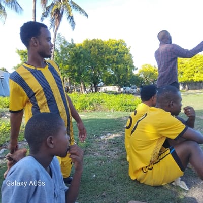 The scene takes place in Nungwi, Tanzania, located at the coordinates -5.726 latitude and 39.301 longitude. The image features four men, three of whom are wearing yellow jerseys with black stripes, sitting on green grass. The men are positioned in a relaxed posture, with the man on the right seeming to be holding a piece of food. The man on the left, wearing a yellow jersey with black stripes, appears to be looking to the left, seemingly engaging in a conversation with someone off-camera. The man standing behind them is wearing a red and black checkered shirt and appears to be looking off-camera. The surrounding environment includes green grass, trees, and buildings.