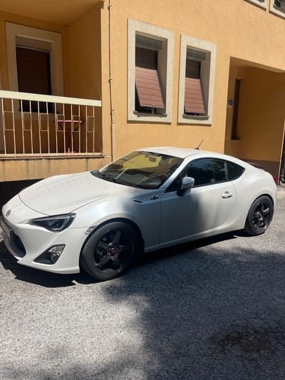 A white, two-door sports car is parked on an asphalt surface outside a building with yellow stucco walls. The car is positioned at an angle, facing towards the left of the frame. It features black alloy wheels with red lug nut caps. The building has several windows with brown shutters and a white railing on a balcony. The scene appears to be set in a residential area, likely a street or courtyard. The lighting suggests it is daytime with strong sunlight casting shadows. The location is specified as Vrela, Montenegro.