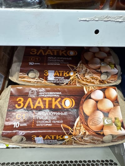 The image shows two cartons of eggs on a shelf, likely in a store. The cartons are brown with a wood-grain pattern and feature the "ЗЛАТКО" (Zlatko) brand name in large yellow letters. The eggs inside are light brown and appear to be standard chicken eggs. Some cartons also display text indicating they are "ЯЙЦА КУРИНЫЕ ПИЩЕВЫЕ СТОЛОВЫЕ ПЕРВОЙ КАТЕГОРИИ" which translates to "CHICKEN EGGS EDIBLE TABLE FIRST CATEGORY". Other labels mention "ОАО «БАРАНОВИЧСКАЯ ПТИЦЕФАБРИКА»" (OAO "Baranovichi Poultry Farm"). One carton has a "TOP" award seal. The setting appears to be a retail environment with a white metal shelf and a honeycomb-patterned metallic surface visible to the right. The lighting is artificial and even. There are no people or animals in the frame. The image is a product shot of breakfast items.