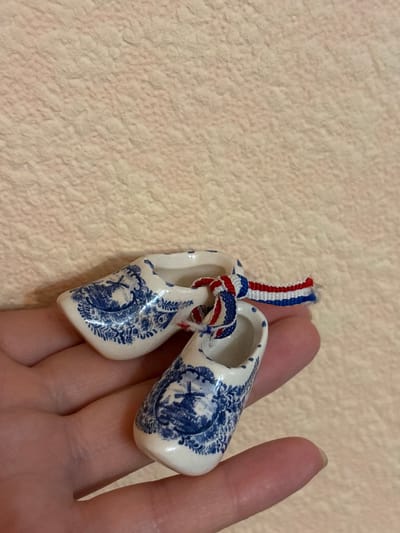 A human hand holds two miniature Dutch clogs decorated with blue windmills and floral patterns. The clogs are tied together with a red, white, and blue ribbon. The background is a textured, light pink surface. The location of Svkh Maslovskiy 1 Otdeleniye, Russia, is not depicted in this image.