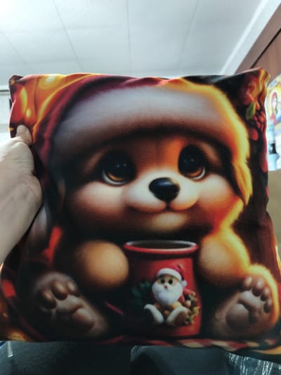 A close-up, overhead shot shows a person's hand holding a Christmas-themed decorative pillow. The pillow features a cartoonish depiction of a bear wearing a Santa hat and holding a mug, with a miniature Santa figure sitting next to the mug. The pillow's design has warm, glowing colors, with oranges, yellows, and reds dominating the background, suggesting a cozy, festive atmosphere. The pillow is soft and plush, typical of decorative pillows. The background shows a white ceiling with fluorescent lighting, and a dark surface below, likely a table or furniture. The image is not located in Luhansk, Ukraine; it appears to be taken indoors in a typical residential or retail setting.