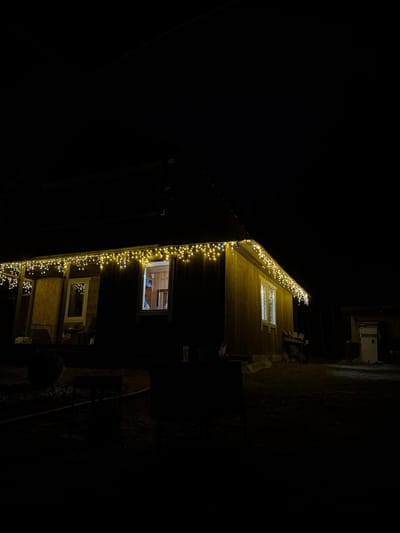 A low-angle shot of a building at night, decorated with string lights. The building's exterior is wooden, with windows illuminated from the interior. The background is dark, with minimal visibility of the surroundings. The location is inferred to be Starosel'e, Russia. No people are visible.