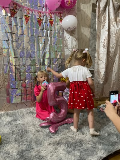 In a room decorated for a child's birthday, two young girls are the focus of the scene. The setting appears to be indoors, possibly a living room, with a fluffy grey rug and a wall adorned with shimmering, reflective squares. A patterned curtain hangs in the background.

The older girl, on the left, is wearing a white t-shirt and a red skirt with white hearts. Her hair is pulled into two pigtails, each adorned with a small white bow. She is facing away from the camera, her back towards the viewer.

The younger girl, on the right, is wearing a bright pink dress and is sitting on the rug. She is holding a pink and blue party blower to her mouth and appears to be blowing into it. She has blonde hair and is looking towards the camera.

Between the two girls are large pink foil balloons shaped like the numbers "5" and "0", suggesting a 50th birthday, though the presence of young children might indicate a party for someone else with these numbers as a theme, or possibly a 5th birthday of one of the children. There are also pink and white balloons scattered around, including one with a Minnie Mouse design and another plain white one. Party banners featuring Minnie Mouse are also visible on the decorated wall.

A hand holding a smartphone is visible in the upper right corner, capturing the moment, indicating that a photo or video is being taken. The overall atmosphere seems festive and joyful, suggesting a birthday celebration. The image is taken in Маринопіль, Ukraine.
