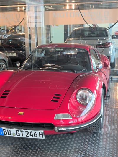 A red Ferrari Dino 246 GT is parked inside a showroom, identifiable by the license plate "BB GT 246 H", which suggests it is registered in Böblingen, Germany. The car is positioned on a textured, diamond-plate floor, likely in a showroom or museum setting. Reflections of overhead lighting and glass are visible on the car's polished surface. In the background, other vehicles are partially visible through the glass, including a grey BMW sedan. Above the cars, a sign with the text "MOTORWORLD" is displayed.