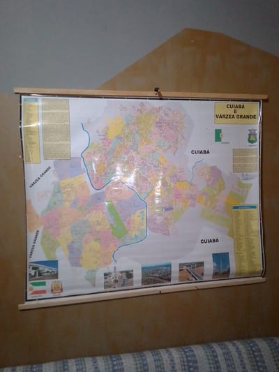 An overhead map of Cuiabá and Várzea Grande, Brazil, is displayed on a wall. The map is bordered by wooden strips at the top and bottom, with a metal rod at the top holding it. Text at the top right of the map reads "CUIABÁ E VÁRZEA GRANDE." The map is detailed, showing streets, neighborhoods, and bodies of water, with various areas color-coded. Several small images are placed along the bottom edge of the map, depicting landmarks, infrastructure, and symbols of the region, including flags and coats of arms. The wall behind the map is a textured, light brown. The bottom of the image shows a portion of a patterned sofa or bed.