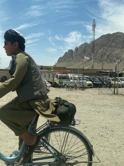 A young Afghan boy is riding a blue bicycle along a dirt road in Arghandab, Afghanistan. He is wearing a traditional Afghan outfit with a vest, head covering, and sunglasses. A black bag is secured to the rear of the bike. In the background, there's a dirt parking area filled with several cars and small trucks. A cell tower stands near a rocky mountain range under a sky filled with wispy clouds. A flag is visible on the mountainside.