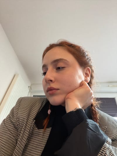 An indoor, close-up image shows a female individual positioned slightly below the camera's viewpoint. She has reddish-brown hair, styled in two visible braids secured with light pink ties. Her left hand supports her chin and jaw. She wears a black long-sleeved top beneath a black and white patterned blazer. A small, light-colored stud earring is visible in her left earlobe, and a silver-colored bracelet is on her left wrist. She looks downwards and slightly to her left. The background consists of a plain white ceiling and upper wall. A portion of a white vertical fixture and a window with closed blinds/shutters are partially visible on the left side of the frame. The image does not contain specific visual cues to confirm the location as Eschau, France.