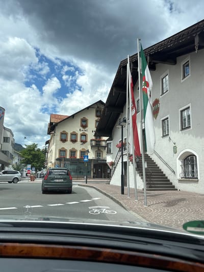 The image captures a street view in Reutte, Austria, on a cloudy day. In the foreground, the dashboard of a vehicle is visible, suggesting the photo was taken from inside a car. The road features painted bicycle lanes and white dashed lines.
A dark green Dacia SUV is in the middle of the road, its rear facing the camera. The car has a license plate that reads "RE 642 64 GA". A sticker on the rear window says "Frühlingsfest Füssen".
To the right of the road, a light-colored building with a dark, ornate roofline is prominent. This building appears to be a hotel and restaurant, with signs for "HOTEL GOLDENER RIEGELER" and "RESTAURANT" visible. Three flags are planted in front of the building: a red and white striped flag, a green and white flag with a crest, and another white flag. An outdoor staircase with a black railing leads up to the building's entrance.
To the left of the hotel, another building with a similar architectural style is seen, also displaying a "HOTEL" sign. Further down the street, more buildings, trees, and street signs are visible. The sky is partly cloudy with dramatic dark clouds in the upper left portion of the image.