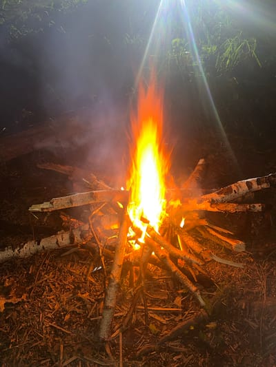 A nighttime scene shows a bonfire. The fire is bright yellow and orange, surrounded by birch logs and other sticks. The area around the fire is dark, with some grass visible in the upper left. There is smoke rising from the fire. The precise location in Krasnyi Jar, Russia, cannot be determined from the image.