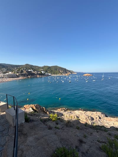 This media file captures a scenic view of a coastal town, likely Tossa de Mar, Spain, from an elevated viewpoint. The scene is dominated by a vast expanse of deep blue water, likely the Mediterranean Sea, stretching out under a clear, bright blue sky. The time of day appears to be midday or early afternoon, given the strong sunlight casting shadows and illuminating the scene.
The coastline is a prominent feature, with a rocky, rugged cliff face on the right side of the image, covered in sparse vegetation. To the left, a densely populated town hugs the hillside, characterized by white buildings with tiled roofs cascading down towards a crowded beach. The beach is a strip of sand packed with many people enjoying the sunny weather.
The water is dotted with numerous boats of various sizes, suggesting a popular recreational area. Several yellow buoys are also visible in the water. The overall atmosphere is one of a vibrant summer day at a seaside resort. The image conveys a sense of leisure and activity, with people visible on the beach and presumably on the boats. The elevated perspective offers a panoramic vista of the town, the sea, and the surrounding natural landscape, highlighting the picturesque setting. There is no visible text in the image.