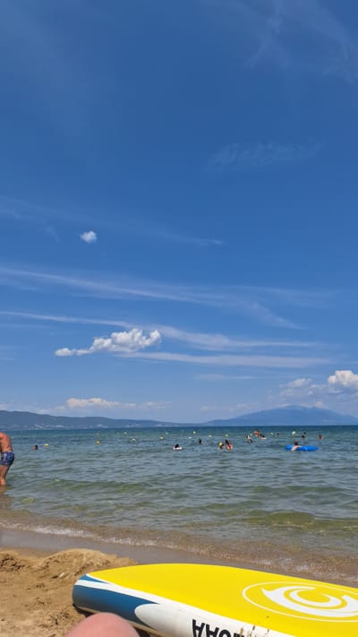The image depicts a sunny day at a beach in Stavros, Greece. The foreground features a yellow and blue inflatable board lying on the sandy shore. The sea is calm and clear, with several people swimming and playing in the water. Yellow buoys mark a designated swimming area. In the background, a range of mountains forms the coastline. The sky is a vibrant blue with scattered white clouds.