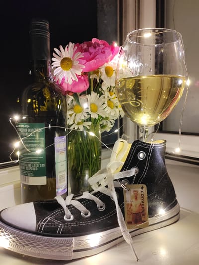 A still life composition is presented, featuring a black Converse sneaker in the foreground, positioned on a white surface. The sneaker has white laces and a small, rectangular tag attached, depicting a vintage photograph of a man in athletic attire with the text "CONVERSE" and "Chuck Taylor" visible. Behind the sneaker, a glass of white wine is prominently displayed, filled to about two-thirds of its capacity and adorned with fairy lights. To the left, a dark green wine bottle stands upright, partially obscured by a bouquet of pink peonies and white daisies in a glass vase. The background is dark and indistinct, suggesting an indoor setting with artificial lighting. The scene is illuminated by fairy lights that are wrapped around the wine glass and the bouquet, casting a warm glow. The image appears to be taken in Nakhodka, Russia, possibly in a domestic setting given the arrangement of everyday items. There are no people present in the image.