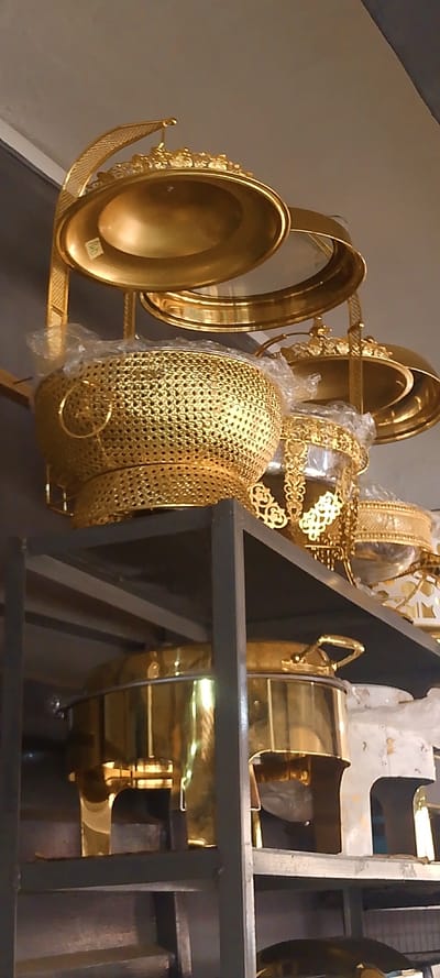 The image displays a collection of golden-colored serving ware and kitchen items arranged on a dark metal shelving unit, located within what appears to be a retail or storage environment in Okere-Urhobo, Nigeria. The upper section of the shelving unit features various decorative golden bowls and serving dishes. One prominent item is a large, oval, golden serving bowl with an ornate, raised rim, exhibiting a small white label on its interior. Directly below it, a golden, perforated bowl with a textured, mesh-like surface and a circular handle on its side is partially encased in clear plastic wrapping. To its right, another golden serving piece with intricate, cut-out scrollwork patterns is also partially covered in clear plastic. The lower shelf holds multiple golden chafing dishes or food warmers. These items are highly reflective, showcasing their polished surface, and feature lids, handles, and robust support structures. The background consists of plain, light-colored walls or ceiling. No individuals are visible in the scene.