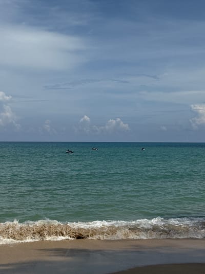 The image depicts a serene ocean scene at Bang Tao Beach in Ban Bang Thao, Thailand. The horizon is clearly defined, separating the tranquil turquoise water from a sky filled with scattered, fluffy white clouds against a backdrop of lighter blue. A gentle wave, tinged with sandy brown sediment, breaks onto the shoreline, which consists of light-colored sand. In the distance, three jet skis are visible on the water. One is positioned to the left of center, with a person on it, while two others are further to the right. The overall impression is one of peaceful coastal recreation.