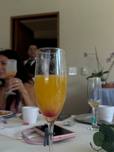 This image captures a close-up of a champagne flute filled with a mimosa, a cocktail typically made with champagne and orange juice. The flute is positioned horizontally in the foreground, obscuring much of the background scene. The drink itself is a vibrant yellow, with a hint of red at the bottom, suggesting the addition of grenadine or a similar ingredient. The flute contains some bubbles, indicating it's freshly poured.
Behind the flute, a table setting is visible, featuring a pink phone with a patterned case, a white cup, and what appears to be a white plate. A plant with green leaves and small purple flowers is also present in the background, adding a touch of natural decor.
The background also reveals reflections and partially obscured figures. A man wearing a suit jacket is visible in a reflection, as is a woman, possibly at a celebration or gathering. The overall lighting suggests it is daytime. The setting appears to be an indoor event, possibly a brunch or party, given the presence of the celebratory drink and formal attire glimpsed in the background. The location context provided, Santiago Yancuitlalpan, Mexico, hints at a potentially festive occasion in this region.