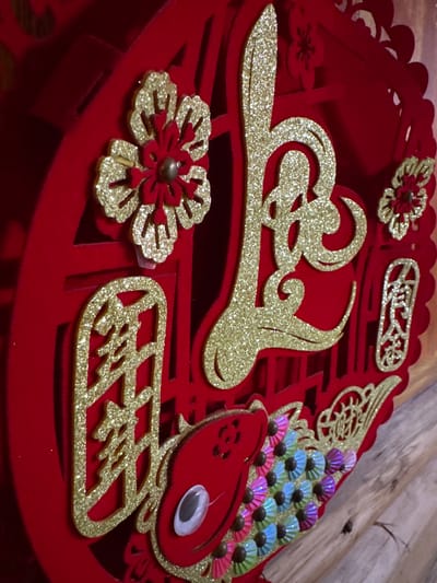 The image features a close-up view of a red, decorative craft object. The object appears to be made of red felt or similar fabric and has intricate cut-out designs. Adorned with glittery gold embellishments shaped like flowers, stylized lettering, and swirling patterns. Notably, there is a red bird-shaped element with what appears to be a googly eye and a fan-like tail made of iridescent, colorful scales. The overall design suggests a celebratory or festive theme. The background is out of focus, showing wooden planks. The location in Mèo Vạc Commune, Vietnam is not discernable from the image alone.