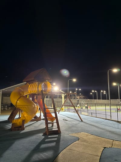 A playground at night. A yellow tunnel slide attached to a wooden structure is illuminated by nearby lights. In the background, a chain-link fence surrounds an area with several people visible under bright lighting. The ground around the playground is a mix of dark, padded material and lighter concrete. Streetlights are positioned above and around the area.