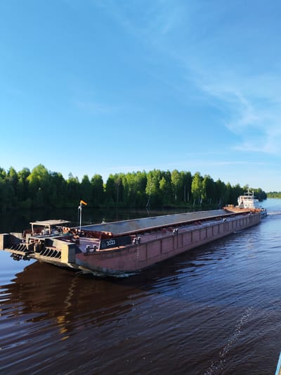 A long, flat-decked barge, likely a cargo vessel, navigates a wide river under a clear blue sky. The barge, predominantly reddish-brown, is moving from left to right across the frame, creating ripples and a wake in the water. Towards the bow (front) of the vessel, a small structure with a light brown canopy and a yellow/orange flag on a pole is visible. The number "3223" is clearly painted in white on a black plate on the side of the barge near the bow.
At the stern (rear), a white cabin or wheelhouse structure can be seen, indicating the vessel's control area. The cargo hold appears empty or covered. Both banks of the river are lined with dense, vibrant green trees, suggesting a lush, natural environment. The sky is bright, with only faint, wispy clouds visible, indicative of a clear, sunny day, possibly morning or late afternoon due to the angle of light on the trees. The water in the river is dark, reflecting the sky and surrounding foliage, with noticeable movement from the barge. The scene depicts a tranquil moment on a Russian waterway, specifically near Annensky Most.