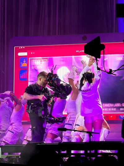 A medium shot captures a live performance, likely at a concert or event. The scene is illuminated by vibrant purple and pink lighting, creating an energetic atmosphere. A large screen in the background displays a digital interface with text such as "KP_LIVE," "CODE:", "2,495,098 watching," and "BAT 85%." Below the screen, a camera operator, dressed in a black shirt and vest, is actively operating a professional video camera rig. He is positioned in front of several performers dressed in white, astronaut-like costumes, some of whom have their arms raised. The performers are on a stage with visible stage equipment and microphones. The image seems to be taken from the audience's perspective. The location in Rivas-Vaciamadrid, Spain, is not discernible from the provided image.