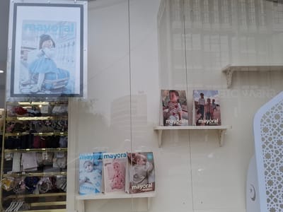 The image displays a store window in Grozny, Russia, showcasing a variety of children's clothing and accessories. Prominently featured are several magazine covers from the brand "Mayoral," which is known for its children's fashion. One large poster on the left depicts a young girl in a blue dress sitting on a suitcase. To the right, on shelves, are three smaller magazine covers featuring children. Below these, on a lower shelf, are three more "Mayoral" magazines with images of babies. A display of children's accessories, including headbands, bows, and other hair ornaments, is visible on the left side of the window, organized on shelves within a glass cabinet. The background of the store window includes reflections of buildings, suggesting an urban environment.