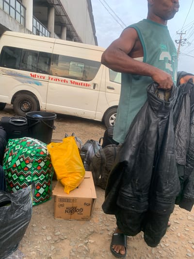A medium shot captures a man in Elelenwo, Nigeria, standing in front of a white shuttle van branded "Silver Travels Shuttle." He is holding several dark-colored jackets, one of which appears to be made of leather. The man wears a sleeveless green shirt and black sandals. The foreground of the image is filled with various bags, including a green and white patterned bag, a bright yellow plastic bag, a black bucket, and several black backpacks and bags. A cardboard box with "GOLDEN MORN" branding is also visible among the luggage. The ground is rough and appears to be dirt or gravel. The background shows a building with a covered walkway and what looks like overhead wires and a utility pole. The sky is overcast.