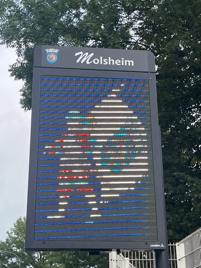 A vertical, black-framed outdoor electronic display board is captured from a slightly low angle against a cloudy sky and dense green tree foliage. The top panel of the board features the word "Molsheim" in white script alongside the Molsheim coat of arms, consisting of a blue shield with an orange and white figure. The main screen of the display exhibits a highly pixelated image, appearing as a fragmented, upward-pointing arrow or conical shape rendered in white and various colors (red, green, blue, yellow) against a background dominated by horizontal blue lines. The display shows visible horizontal banding and apparent pixel or panel malfunction, presenting a striated pattern across the image. At the bottom right corner of the display, the text "Lumiplan" and a stylized human figure logo are visible. To the bottom right of the sign, a light-colored metal fence or railing is partially visible. The location indicated on the sign is Molsheim, France.