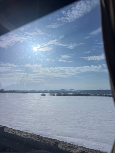 The image displays a winter landscape under a bright, clear blue sky with scattered white cumulus clouds, captured from inside a vehicle. The sun, a prominent luminous circle, is positioned in the upper-left quadrant, generating significant lens flare and glare across the upper half of the view. The foreground, partially obscured by the vehicle's interior elements, shows a dark, textured roadside embankment or barrier. Extending from the midground to the horizon is a vast, flat field covered uniformly in white snow, with sparse dark patches of vegetation visible. A distinct treeline of leafless deciduous trees forms a band across the middle distance, beyond which gently rolling, snow-covered hills are discernible. No specific individuals, animals, or structures are visible within the natural landscape. The scene is located in Chernovskoe, Russia.