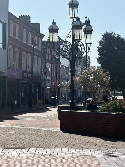 This image depicts a pedestrianized section of Market Street in Crewe, United Kingdom, under bright sunlight. In the foreground, a prominent black, ornate metal lamp post with five lantern heads stands centrally; one top lantern shows a damaged or missing panel. To the immediate right, a reddish-brown brick planter box contains green benches and foliage. A person wearing a dark top is seated on one of these benches, facing away from the viewer. The pavement consists of reddish-brown and grey brick-style pavers, interspersed with lighter tactile strips.
On the left side of the street, a row of multi-story commercial buildings is visible. The closest visible businesses at ground level are "Vision Express" and "HUFF & PUFF," the latter featuring a yellow A-frame sign outside. Further down the street, various architectural styles are present, including structures with arched windows and some half-timbered facades.
In the midground and background, the street continues, lined by more buildings and shops. Several pedestrians are discernible on the pavement, moving or standing. A magnolia tree with white blossoms is prominent in the midground on the right, adjacent to a larger, dark green evergreen tree. The scene is illuminated by strong sunlight, casting distinct shadows across the pavement and buildings.