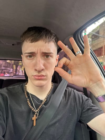 An individual is depicted in a close-up, front-facing selfie taken within the interior of a vehicle in Caseros, Argentina. The subject, appearing male, has short brown hair, fair complexion, and brown eyes. A silver stud piercing is present in the left nostril, a silver labret piercing below the lower lip, and a small silver hoop earring in the left earlobe. The subject wears a dark-colored t-shirt. Around the neck are two necklaces: an outer necklace of brown wooden beads with a light brown wooden cross pendant, and an inner thin silver chain. A dark-colored seatbelt diagonally crosses the subject's chest. On the fourth finger of the right hand is a silver-toned band ring. A wristwatch with a purple strap and silver-toned casing is worn on the left wrist. The subject's right hand is raised to the right of the face, forming an "OK" gesture with the thumb and index finger connected, and the remaining three fingers extended vertically. The subject maintains eye contact with the camera, exhibiting a neutral facial expression with a slightly downturned mouth. The vehicle's grey headliner is visible at the top of the frame, and parts of the car's interior frame are visible on the right. Through the vehicle's windows, a blurred urban street scene with buildings, trees, and overhead wires is visible, indicating an outdoor setting during daylight hours.