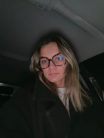 A woman with blonde hair and glasses is angled diagonally in the frame. She wears a dark coat or jacket, and a white shirt peeks out at the collar. The background appears to be the interior of a car, with dark and light grey surfaces visible. The lighting is dim, creating shadows around her face. The reflection of a screen is visible in her glasses.