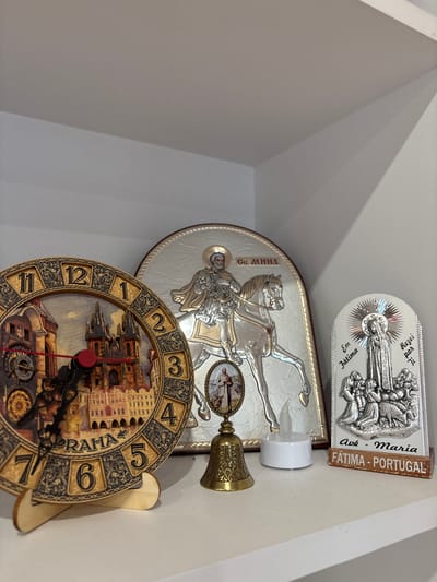 This image shows a collection of religious and decorative items placed on a shelf or ledge against a white, angled wall. The subjects are primarily religious artifacts and a clock.
In the foreground, a round clock with an ornate, antique design is visible. The clock face features a detailed illustration of buildings, likely a cityscape, and the numbers are stylized and embedded within decorative elements. The words "PRAHA" are clearly visible along the right side of the clock face, indicating it is a Prague-themed clock. The clock has black hour and minute hands, with a red second hand.
Above the clock is a silver oval plaque depicting Saint George on horseback, slaying a dragon. The saint is rendered in intricate detail, and the plaque has text in Cyrillic script, "Св. Мина" which translates to "St. Mina". There is also a small, ornate brass bell with a handle attached to a decorative element. Next to the bell is a small, white, LED candle with a flickering flame effect.
Further up, another silver oval plaque is visible. This plaque features a religious scene with figures, and text in Portuguese that reads "Ave Maria", "Em Fátima", and "Rezei por Ji". The bottom of this plaque has a wooden strip with text "FÁTIMA - PORTUGAL".
The setting appears to be indoors, possibly a religious or decorative display. The lighting is bright, suggesting daytime. There are no people visible in the image. The overall impression is one of personal devotion or a collection of souvenirs.