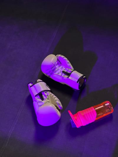 This media file captures a still life composition of boxing gloves and a water bottle, set against a dark, possibly fabric, surface. The scene is illuminated by purple and pink lighting, creating a dramatic and moody ambiance. Two white boxing gloves with "EVERLAST" text printed repeatedly on them are positioned on the left and lower right of the frame. Between them, a bright pink and orange water bottle with a flexible grip and a flip-top cap rests at an angle. The lighting casts long shadows, suggesting a low light environment or artificial lighting specifically designed for effect. The overall impression is one of preparation for a workout or a post-workout scene, with an emphasis on athletic gear and hydration. There are no people or animals visible. The visible text on the gloves clearly reads "EVERLAST". The location context provided is Kâğıthane, Turkey, but there are no specific visual cues in the image to confirm this location.