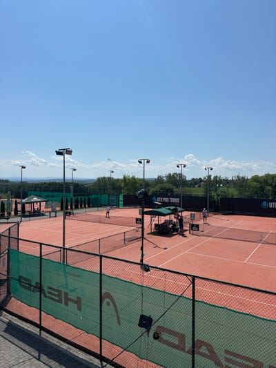 The image depicts an outdoor tennis complex, identified as the Sport Klub Tenisowy Giebułtów, located at ul. Spacerowa 1, 32-085 Giebułtów, Poland. The scene is illuminated by bright daylight under a clear blue sky with scattered cumulus clouds.
In the foreground, a dark green chain-link fence extends across the bottom of the frame, partially covered by a green windbreak bearing the inverted white "HEAD" logo. Attached to this fence are two dark-colored, vertical poles with mounted equipment, possibly for broadcasting or recording. A paved or tiled walkway is visible at the lower left.
The midground features multiple red clay tennis courts with white boundary lines. At least four courts are discernible. On the leftmost visible court, two individuals are actively engaged in play; one player, dressed in white, is positioned near the baseline. On the rightmost visible court, two additional individuals are present, with one nearer the net and the other further back. Between these two courts, a green canopy tent provides shade, under which at least two individuals are gathered around a table with various objects, including coolers. A tall, dark light pole with a large rectangular fixture stands prominently near this canopy, equipped with additional mounted devices.
The facility is equipped with numerous tall, grey light poles, each topped with a dark rectangular light fixture, positioned strategically around and between the courts.
In the background, dark perimeter fencing and privacy screens line the courts. Black banners displaying "UTR PRO TENNIS TOUR" in white text are visible along the far side of several courts. Beyond the immediate court area, dense green trees and vegetation form a natural backdrop. In the distant background, rolling hills or elevated terrain are visible under the horizon line, indicative of a rural or semi-rural landscape.