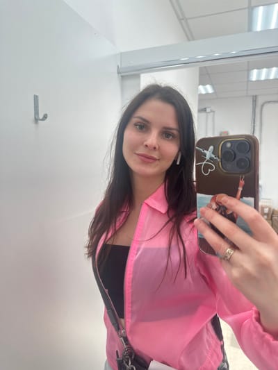 A young woman with long dark hair and green eyes is taking a selfie in a room with white walls and fluorescent lighting. She is wearing a black top and a sheer pink jacket. She has white wireless earbuds in her ears and is holding a smartphone with a decorative case. A ring is visible on her right hand. The room appears to be an indoor space with a hook on the wall and some boxes in the background. The image is likely taken in the city of Novoselye, Russia.