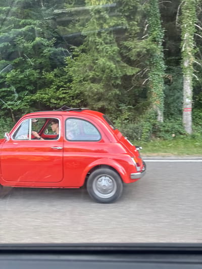 A red Fiat 500 is driving on a paved road surrounded by lush green trees and vegetation. The car, seen from the side, has its sunroof open. A person wearing glasses and a dark t-shirt is visible driving the car. The background consists of dense woods with ivy growing on some of the trees. The image appears to be taken from a moving vehicle, as indicated by the motion blur on the road. The location is the commune of Villards-d'Héria, France.