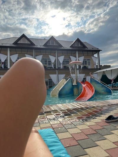 In the city of Yaroslavskaya, Russia, a poolside scene unfolds under a cloudy sky. The foreground is dominated by a blurred close-up of a person's leg and thigh, resting on a turquoise cushion. To the right, a section of the poolside paving is visible, composed of rectangular tiles in various earthy tones. A single navy blue Croc shoe rests on the tiles near the water's edge.
Beyond the paving lies a shallow pool with clear blue water. A children's water slide structure is the central feature of the pool area. The slide is composed of blue and red plastic sections, with a yellow slide on the left and a bright orange slide on the right curving into the water. A mushroom-shaped water feature is positioned above the slides, showering water down.
In the background, a two-story building with a dark, corrugated metal roof stands behind the pool. The building features multiple balconies with white curtains draped across them, suggesting hotel rooms or apartments. The overall atmosphere is one of leisure and relaxation, despite the overcast sky. A young child can be seen standing on the steps leading up to the water slide.
