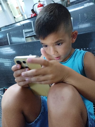 A young boy, appearing to be around 8-10 years old, is seated and focused on a golden smartphone held in his hands. He is wearing a blue sleeveless shirt and denim shorts. The boy's hair is dark and cut short with a distinct fade on the sides. He is looking down intently at the phone, his fingers manipulating the screen. In the background, a row of dark, perforated seats suggests a waiting area, possibly in a public transport station or a similar facility. A fire extinguisher is visible mounted on a wall further back. Faint writing is scrawled on the seat in front of the boy, too indistinct to be fully deciphered but appearing to contain letters and symbols. The lighting is diffused, indicating an indoor environment. The overall scene suggests a moment of quiet engagement with technology in a public space.