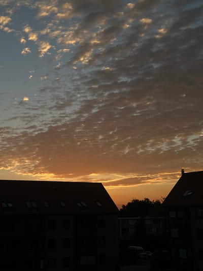 The image captures a sky filled with fragmented, low-lying clouds illuminated by the warm hues of a sunset or sunrise. The sky transitions from a lighter, cooler blue at the top to a vibrant orange near the horizon. Silhouetted against this colorful sky are the dark shapes of buildings, their red-tiled roofs and windows barely discernible. The architecture appears to be residential, with multiple stories and dormer windows visible on one of the structures. Trees form a dark band between the sky and the lower buildings. The scene is set in the city of Roskilde, Denmark.