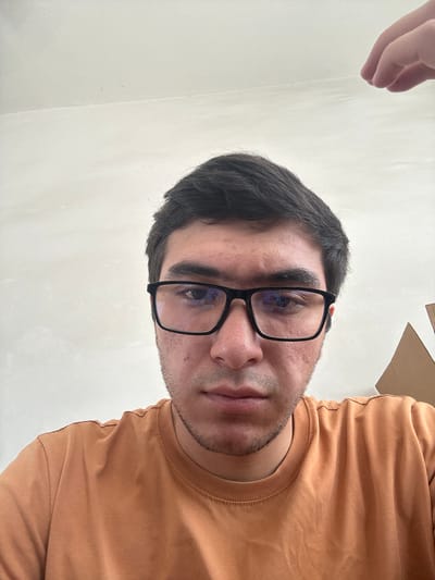 This is a close-up, waist-up, selfie-style portrait of a young man with dark hair and a light complexion, wearing black-rimmed glasses and an orange t-shirt. He has a neutral expression and is looking directly at the camera. The background appears to be an indoor environment with a plain, light-colored wall. There is also a partial view of a cardboard box to the right of the man, and a hand, out of focus, is visible in the upper right corner of the image, suggesting someone else is present. The image does not contain any information about the city of Danghara, Uzbekistan.