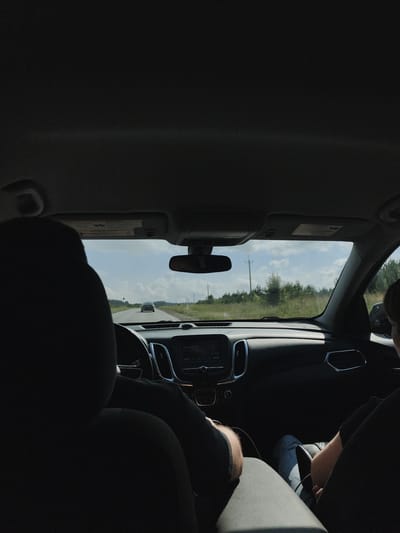 The image shows the interior of a car looking forward through the windshield. The foreground is dominated by the dark silhouettes of two car seats, suggesting the perspective of someone in the back seat. The dashboard of the car is visible, with a screen displaying "Just Lose It". Outside the car, a paved road stretches into the distance, lined with grassy fields and trees. A light-colored car is driving ahead on the road, and power lines are visible in the background. The sky is partly cloudy. The image appears to have been taken during the daytime. There is no information in the image to indicate the location is in Dokshitsy, Russia.